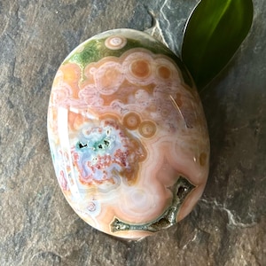 May include: Polished oval gemstone featuring a swirling pattern of peach, cream, green, and blue. The stone has a glossy finish and a natural appearance. The gemstone is a decorative item.