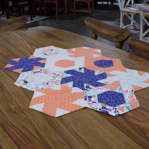 Könnte beinhalten: Ein sechseckiger Tischläufer mit Patchwork-Design aus blauem, orangefarbenem und weißem Stoff. Der Läufer besteht aus verschiedenen geometrischen Formen und floralen Mustern.