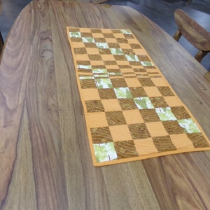 May include: A checkered table runner with a brown and green pattern. The runner is made of fabric and has a quilted texture. It is laid out on a wooden table.