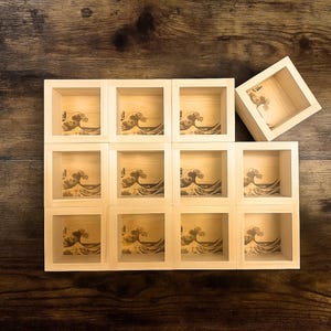 May include: Wooden sake boxes with a wave design. The boxes are arranged in a grid, with one box slightly off to the side. The wave design is etched into the wood, creating a detailed and artistic look. The boxes are made of light-colored wood.
