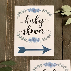 May include: Two white signs with blue floral wreaths and arrows pointing to the right. The signs say "baby shower" in black cursive.