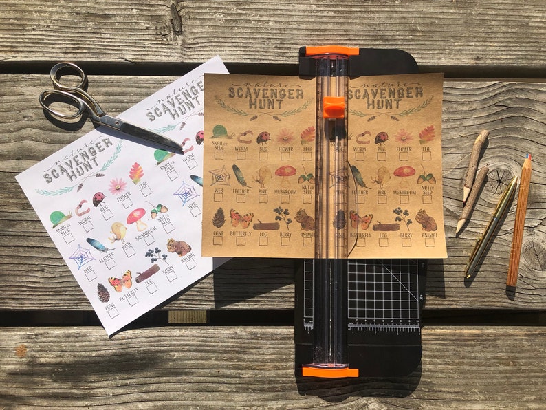 May include: Two printable nature scavenger hunt checklists. One checklist is printed on white paper and the other is printed on brown paper. The checklists feature illustrations of various nature items, such as a ladybug, a flower, a feather, a mushroom, a butterfly, a pine cone, and a berry. The checklists are laid out on a wooden surface with a pair of scissors, a pencil, and a craft knife.