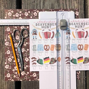 Pu&ograve; includere: Due carte di gioco di caccia al tesoro di Oktoberfest stampabili con un elenco di controllo degli oggetti da trovare, tra cui lederhosen, un pretzel e una fisarmonica. Le carte sono su uno sfondo bianco con linee di griglia marroni. Le carte sono su una superficie di legno con un righello, delle forbici e una matita.