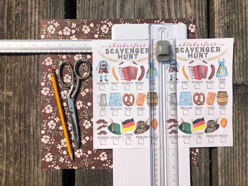 Pu&ograve; includere: Due carte di gioco di caccia al tesoro di Oktoberfest stampabili con un elenco di controllo degli oggetti da trovare, tra cui lederhosen, un pretzel e una fisarmonica. Le carte sono su uno sfondo bianco con linee di griglia marroni. Le carte sono su una superficie di legno con un righello, delle forbici e una matita.