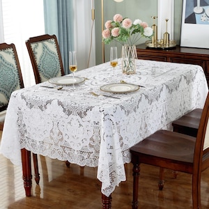 Puede incluir: Un mantel de encaje blanco cubre una mesa de comedor de madera, con platos, vasos y cubiertos. El intrincado diseño de encaje es visible, y la mesa está rodeada de sillas de madera. Un jarrón con flores está en el centro.