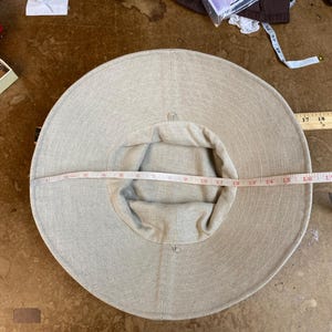 May include: A beige sun hat with a wide brim, approximately 40 cm in diameter. The hat is made of a light-colored fabric and has a rounded crown. A measuring tape is placed across the hat to measure its diameter.
