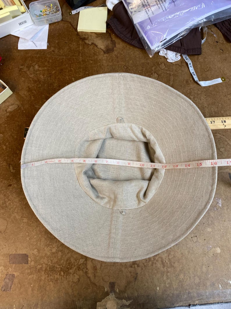 May include: A beige sun hat with a wide brim, approximately 40 cm in diameter. The hat is made of a light-colored fabric and has a rounded crown. A measuring tape is placed across the hat to measure its diameter.