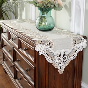 May include: A decorative beige and white lace table runner with a floral design is displayed on a dark brown wooden dresser. A green glass vase with flowers sits on top of the dresser.