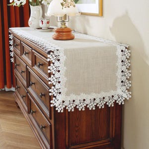 May include: A beige linen table runner with white crocheted lace trim. The runner is on a wooden dresser with drawers.