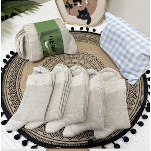 Könnte beinhalten: Mehrere Paar hellbeige Socken, einige ordentlich gefaltet und mit einem grünen Band mit der Aufschrift "HEMP" umwickelt. Die Socken haben ein helles Zehen- und Fersendesign. Ein blau-weiß kariertes Kissen befindet sich im Hintergrund.