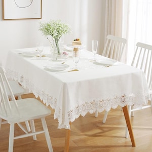 May include: A rectangular white tablecloth featuring an intricate lace border with scalloped edges. The fabric is smooth and opaque, draped over a dining table set with white plates, clear glassware, and a glass vase filled with white flowers.