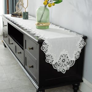 Könnte beinhalten: Ein weißer Tischläufer aus Spitze mit floralem Design liegt auf einem schwarzen Schrank. Der Läufer hat einen gewellten Rand und ist aus zartem Stoff gefertigt. Eine grüne Vase mit gelben Früchten steht auf dem Schrank.
