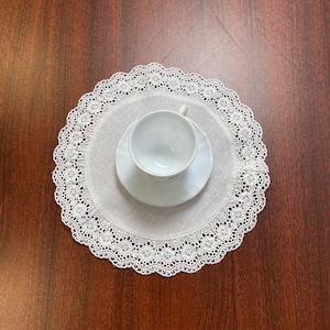 Könnte beinhalten: Eine weiße Teetasse und Untertasse auf einer weißen Spitzendecke. Die Deckchen hat einen gewellten Rand mit einem detaillierten Blumenmuster. Die Gegenstände sind auf einer dunkelbraunen Holzoberfläche angeordnet.