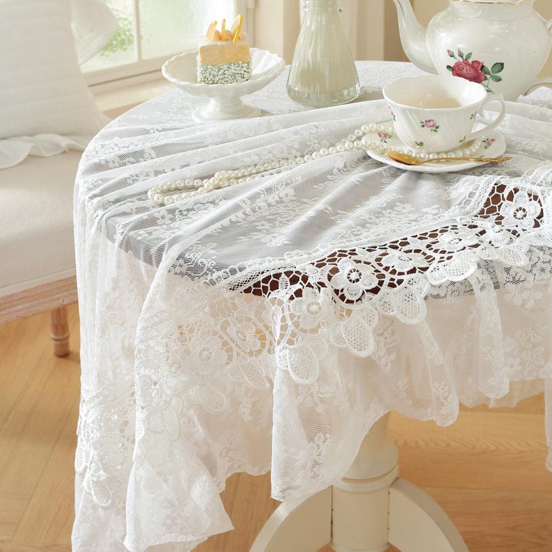 K&ouml;nnte beinhalten: Eine wei&szlig;e Spitzentischdecke mit gewelltem Rand bedeckt einen runden Tisch. Eine Tasse und Untertasse, eine Teekanne mit Rosenmuster und eine Perlenkette liegen auf dem Tisch. Ein kleiner Kuchen steht auf einem Fu&szlig;teller.
