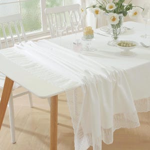 May include: A white table setting with a white tablecloth and a decorative white lace table runner. The table is set with white chairs, glassware, and a vase of white flowers. A small cake and decorative bowl are also on the table.