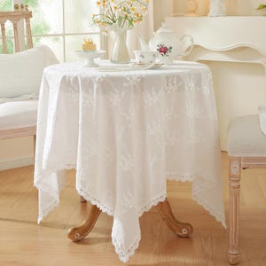 Puede incluir: Un mantel redondo de encaje blanco con un diseño floral cubre una pequeña mesa de madera. Una tetera, una taza de té y un soporte para pasteles están colocados sobre la mesa, creando una estética vintage.