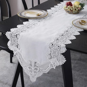 May include: A white lace table runner featuring detailed floral and scroll patterns. The runner is laid on a black table, with plates and a fruit bowl in view. The lace trim provides an elegant touch.