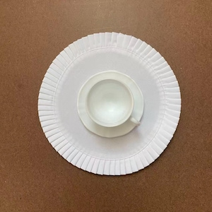 May include: A white teacup and saucer on a white, circular placemat with a pleated border. The teacup is positioned centrally on the saucer, which rests on the placemat. The placemat is on a brown surface. A simple, clean design.