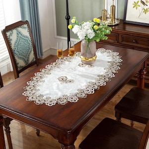 May include: A white and gold embroidered table runner with a floral design is displayed on a dark wood dining table. The runner has a delicate lace trim. Two champagne flutes and a vase of flowers are on the table.