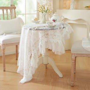 May include: A small, round table covered with a white lace tablecloth. The lace has detailed floral designs and a ruffled trim. A white teapot with a rose motif and a teacup are on the table, with a vase of flowers adding to the elegant setting.