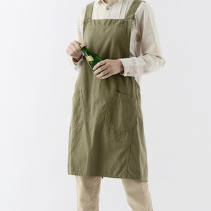 May include: An olive green apron with two large front pockets and cross-back straps. The apron is made of a textured fabric. The person is holding a green bottle. The apron has a simple, functional design.