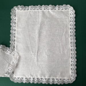 May include: A rectangular white linen placemat featuring a delicate, intricate white lace border along all four edges. The fabric has a visible woven texture, and a corner of a matching second placemat is partially visible on the left side.