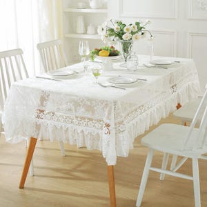 May include: White lace tablecloth with a ruffled edge, set on a wooden table with four chairs. The tablecloth has a delicate floral pattern and is perfect for a formal dining setting.