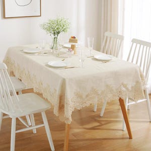 以下が含まれることがあります： 繊細なレースの縁取りが施されたベージュのテーブルクロス。テーブルクロスは白い皿、カトラリー、グラスが置かれています。テーブルには白いケーキが載った小さなケーキスタンドがあります。テーブルは白い椅子で囲まれています。