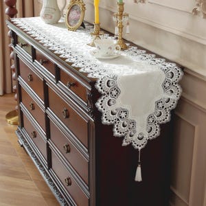 Puede incluir: Camino de mesa de encaje blanco con un diseño floral detallado y una borla. El camino de mesa se exhibe sobre una cómoda de madera marrón oscuro, con elementos decorativos como una taza de té y candelabros.
