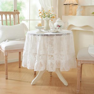 May include: A small, round white table covered with a white lace tablecloth. The table is set for tea, with a teapot, teacup, and a vase of flowers. A white chair is next to the table.