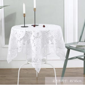 Puede incluir: Un mantel de encaje blanco, de aproximadamente 85 cm por 85 cm, cubre una pequeña mesa redonda. Dos candelabros negros con detalles dorados sostienen velas blancas. Una silla verde claro está en el fondo.