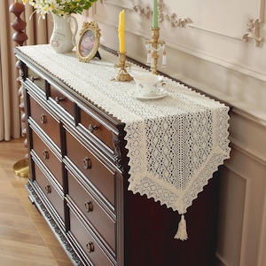 May include: Cream-colored lace table runner with a scalloped edge and tassel. The runner is placed on a dark brown wooden dresser, with decorative items like candles, a picture frame, and a teacup.