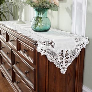Puede incluir: Un camino de mesa de encaje blanco con un intrincado diseño floral se muestra sobre una cómoda de madera marrón oscuro. Un jarrón de cristal verde con flores está encima.