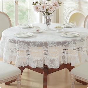May include: A round dining table covered with a white lace tablecloth featuring a floral pattern and a ruffled edge. The table is set with white plates, silverware, clear glasses, a small bowl, a teacup, and a glass vase with pink flowers.
