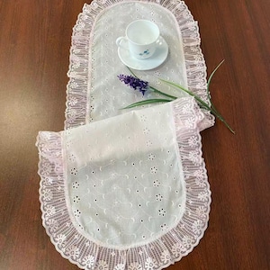 Puede incluir: Camino de mesa de encaje blanco con borde rosa con volantes. Una taza de té y un platillo blancos con un diseño floral se encuentran en el camino de mesa, junto con una ramita de flores moradas. El camino de mesa está sobre una superficie de madera marrón oscuro.