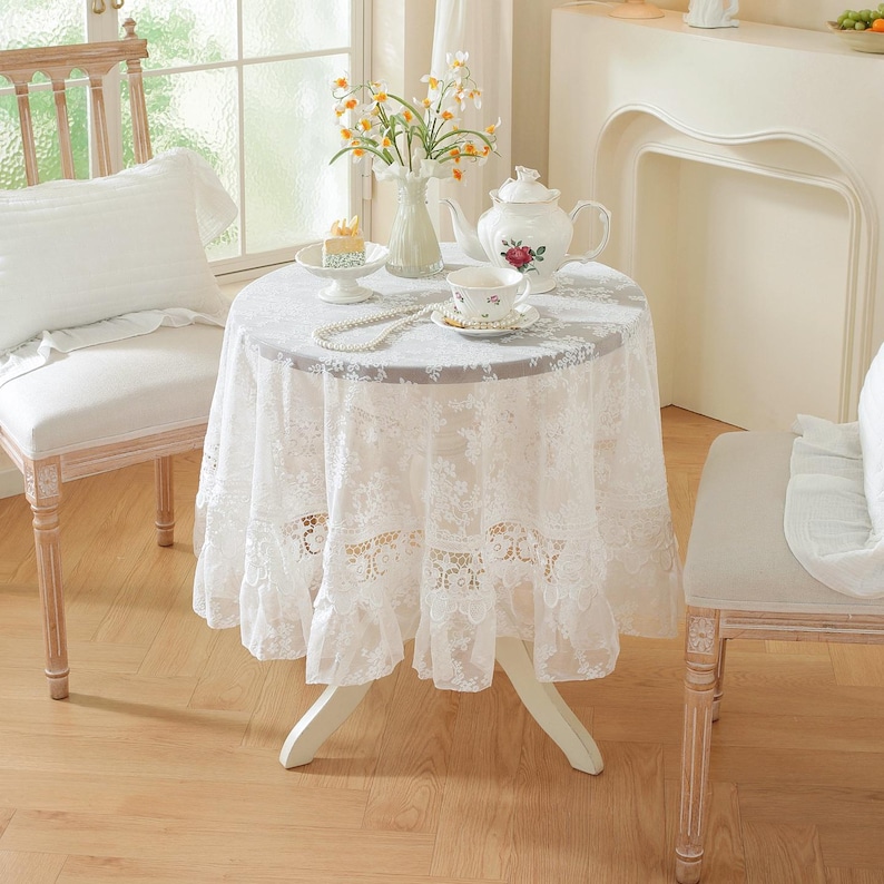 K&ouml;nnte beinhalten: Ein kleiner runder Tisch, bedeckt mit einer wei&szlig;en Spitzentischdecke. Auf dem Tisch befindet sich ein wei&szlig;es Porzellan-Teeservice mit floralem Design, zusammen mit einem kleinen Kuchen und einer Blumenvase. Zwei St&uuml;hle flankieren den Tisch.