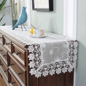 May include: A white lace table runner with floral and leaf patterns is displayed on a dark wood dresser. The runner has a delicate, openwork design and is placed under a blue bird statue and a teacup.