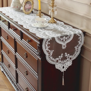 Può includere: Una tovaglia in pizzo bianco con ricami floreali è esposta su una cassettiera marrone scuro. La tovaglia ha un bordo smerlato e un nappa. Una tazza da tè, candelabri e una cornice sono sulla cassettiera.