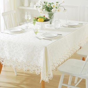 Puede incluir: Mantel de encaje blanco con un delicado patrón floral, preparado para una cena formal con platos blancos, cubiertos y un jarrón de flores blancas.