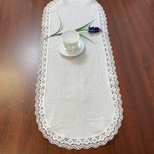 May include: A white oval table runner with a delicate lace trim. A white teacup and saucer sit on the runner, along with a sprig of lavender. The runner is placed on a dark wood surface.