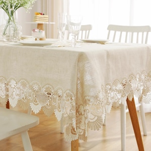 May include: A beige rectangular tablecloth with a decorative white lace border. The tablecloth is set on a wooden table with white chairs. The table is set with plates, glasses, and a cake stand.