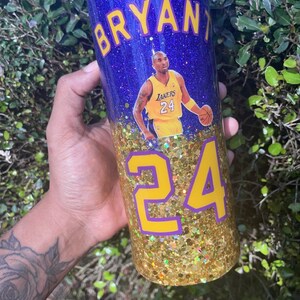 May include: A purple glitter tumbler with a gold glitter bottom. The tumbler has a picture of Kobe Bryant in a Lakers jersey with the number 24 on it. The text "BRYANT" is on the top of the tumbler and the number "24" is on the bottom.