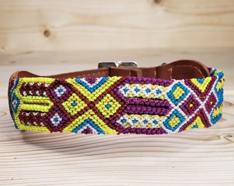 May include: A colorful dog collar with a geometric pattern in shades of yellow, purple, blue, and red. The collar is made of woven string and has a brown leather backing.
