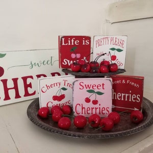 May include: A set of six wooden signs with red and white paint and cherry graphics. The signs say "Life is sweet", "Pretty please with a cherry on top", "Cherry Tree Cottage", "Sweet Cherries", "Sweet & Fresh", and "Cherries". The signs are arranged on a tiered stand with a silver tray of red cherries.