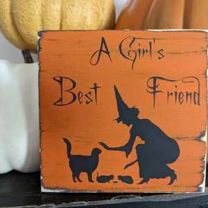 May include: Orange wooden sign with the text "A Girl's Best Friend" and a black silhouette of a witch and cat. The sign has a distressed, rustic look, perfect for Halloween decor.