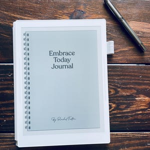 Può includere: Un diario bianco con il testo "Embrace Today Journal" e una rilegatura a spirale argentata. Una penna d'argento è appoggiata su una superficie di legno scuro accanto al diario. Il diario ha una copertina grigio chiaro.