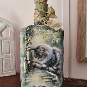 May include: Decorative bottle with a detailed painting of a fluffy gray cat resting on a tree branch above a pond with a red fish. The bottle's neck is adorned with a jeweled stopper and camouflage ribbon, and a beaded chain hangs from the neck.