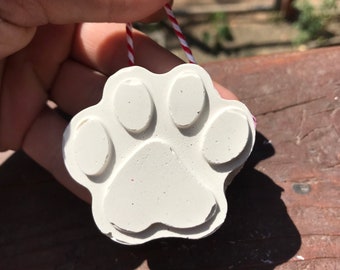 May include: A white paw print ornament with a red and white string. The ornament is held by a hand with pink nail polish. The ornament is made of a solid material and has a detailed paw print design.