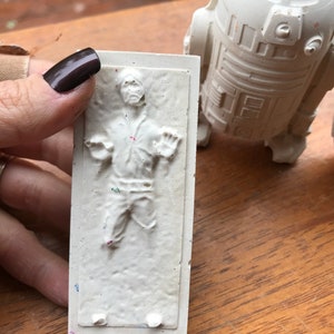 K&ouml;nnte beinhalten: Eine wei&szlig;e Reliefskulptur einer Figur in einer gefrorenen Pose, mit einer passenden wei&szlig;en R2-D2-Figur im Hintergrund. Die Skulptur wird von einer Hand mit dunklem Nagellack gehalten. Die Figuren befinden sich auf einer Holzoberfl&auml;che.