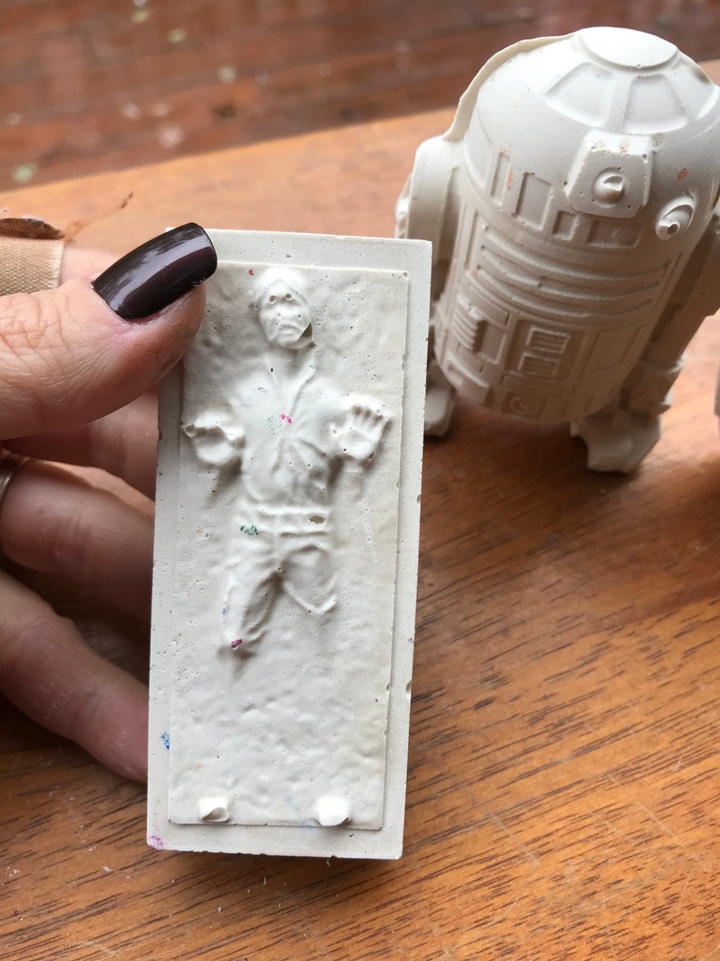 K&ouml;nnte beinhalten: Eine wei&szlig;e Reliefskulptur einer Figur in einer gefrorenen Pose, mit einer passenden wei&szlig;en R2-D2-Figur im Hintergrund. Die Skulptur wird von einer Hand mit dunklem Nagellack gehalten. Die Figuren befinden sich auf einer Holzoberfl&auml;che.